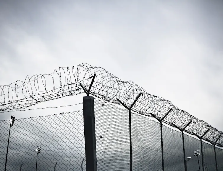 A grayscale view of a tall chain-link prison fence topped with rows of barbed wire