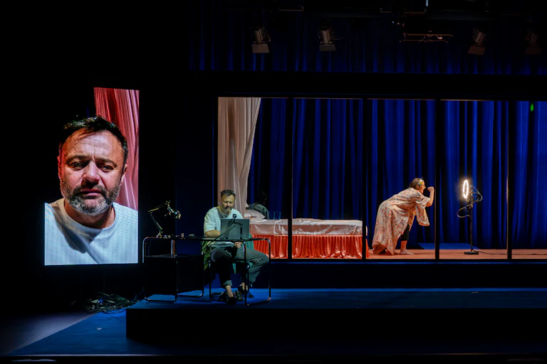 Production image: a man sits at a laptop, his large face projected behind.