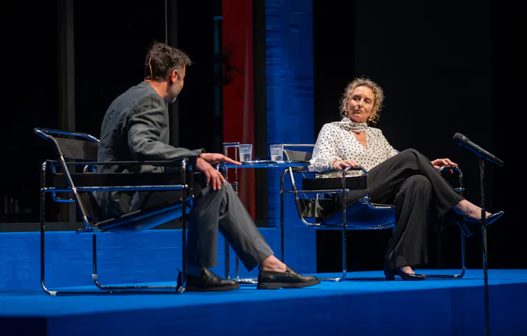 Production image: a man and a woman talk on a panel.