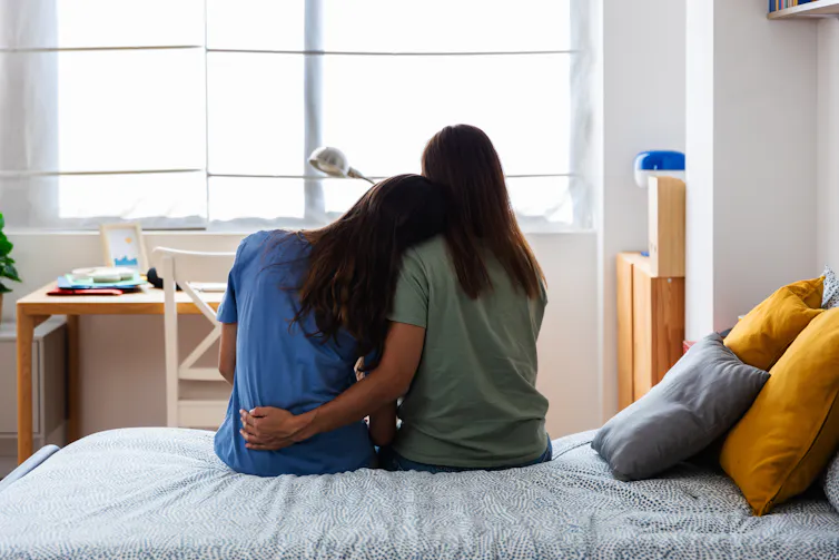 view from behind of a mother and daughter embracing while sitting on a bed at home
