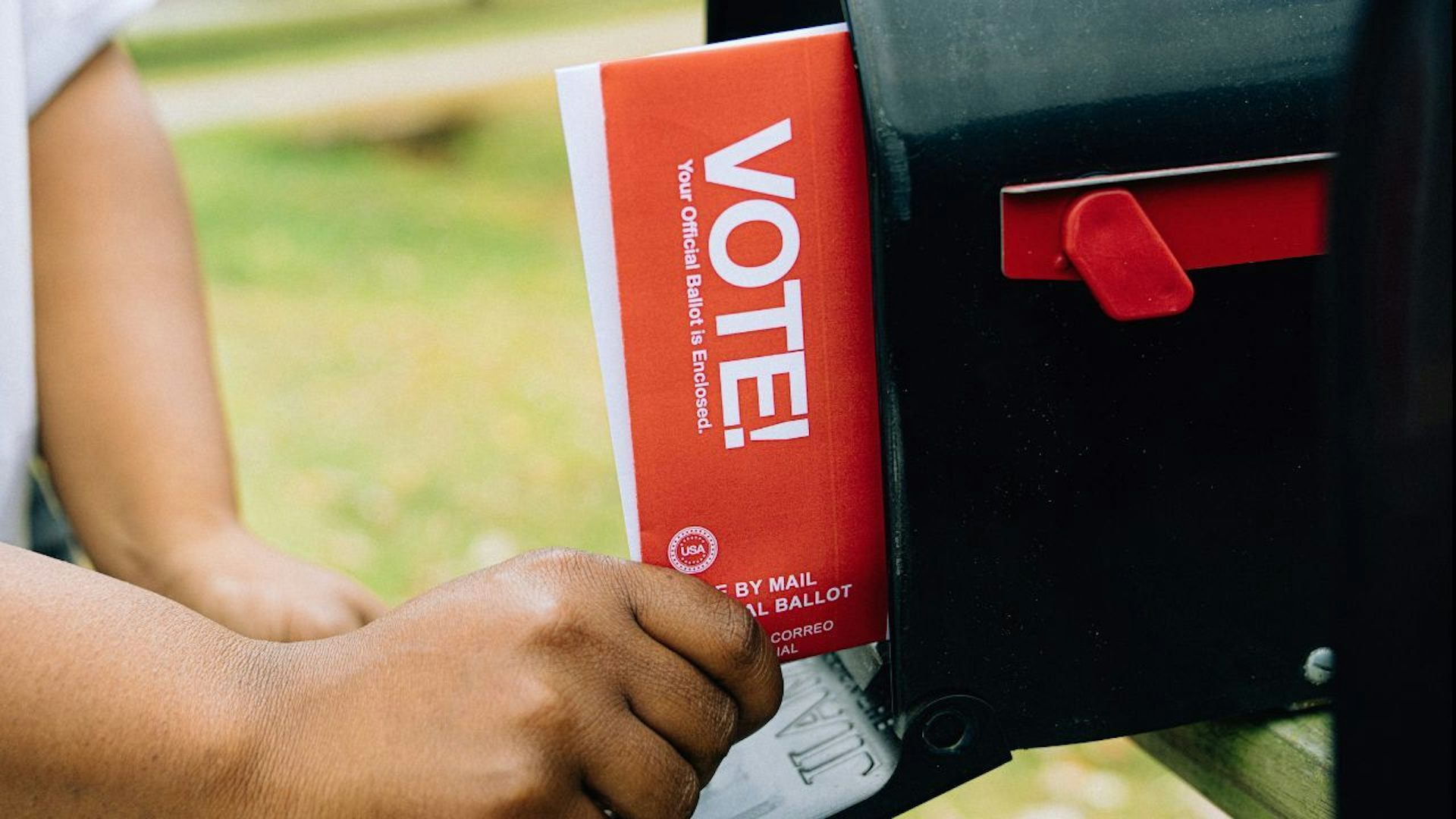 person puts a ballot in a black mailbox that says 'vote!' on it in white lettering on a red background