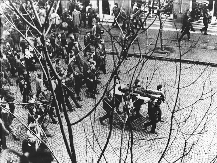 Fotografía en blanco y negro de un grupo de manifestantes que siguen a un grupo que lleva el cadáver de un hombre sobre una puerta.