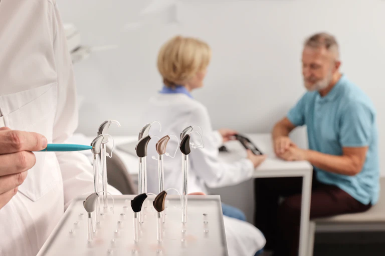 Au premier plan, un professionnel de santé montre un présentoir avec différentes prothèses auditives, pendant qu'une personne âgée au second plan est en consultation avec une autre professionnelle.