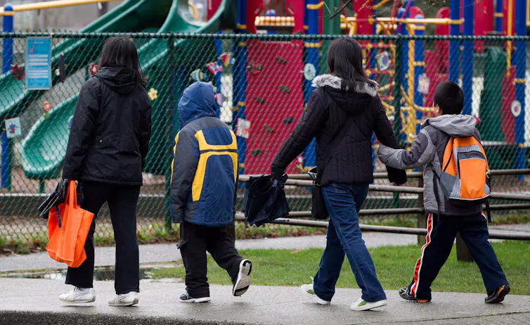 The backs of people seen walking near a playground fence outdoors.