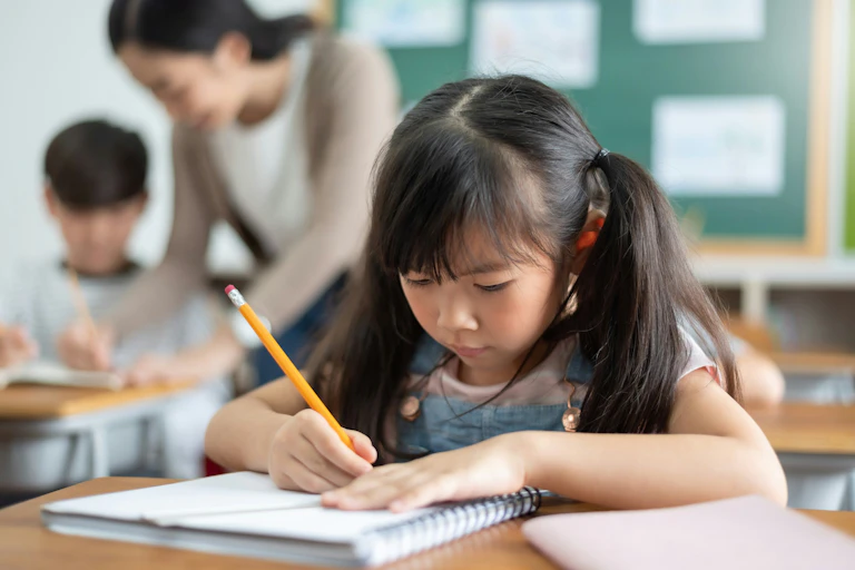 An Asian child in a classroom writing in a notebook with a pencil and behind her is a teacher in blurred focus with a student.