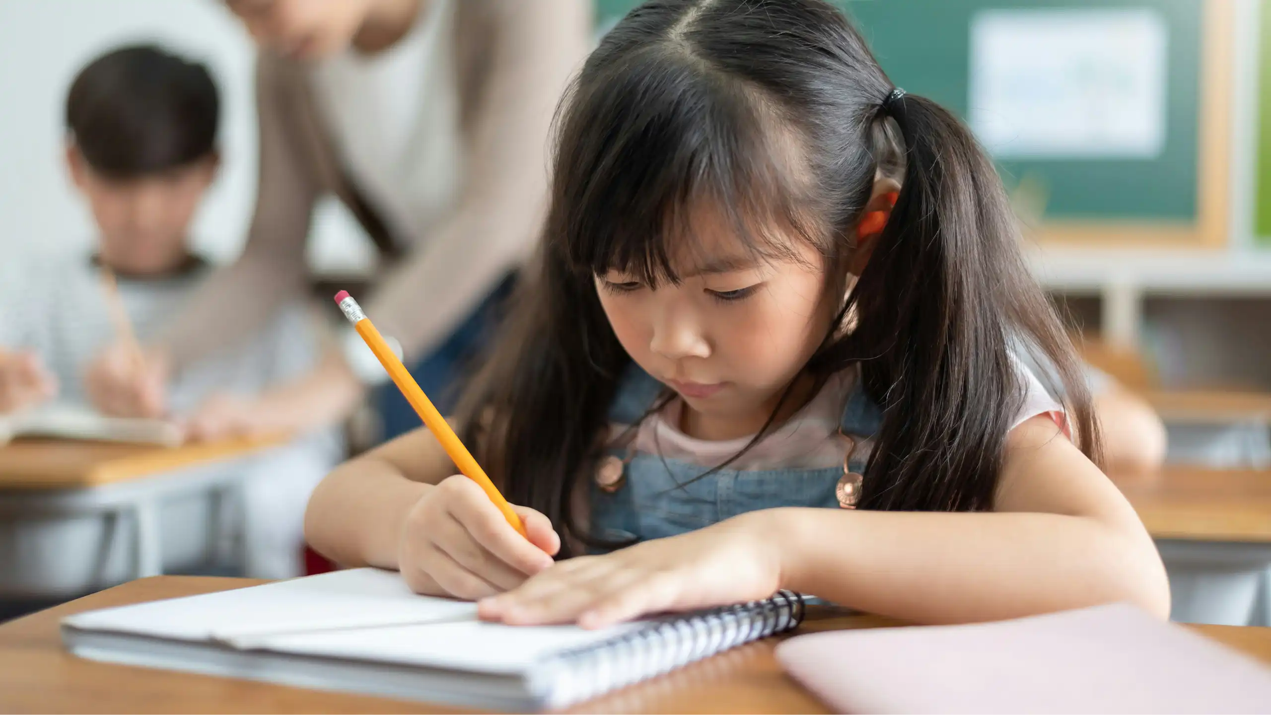 An Asian child in a classroom writing in a notebook with a pencil and behind her is a teacher in blurred focus with a student.