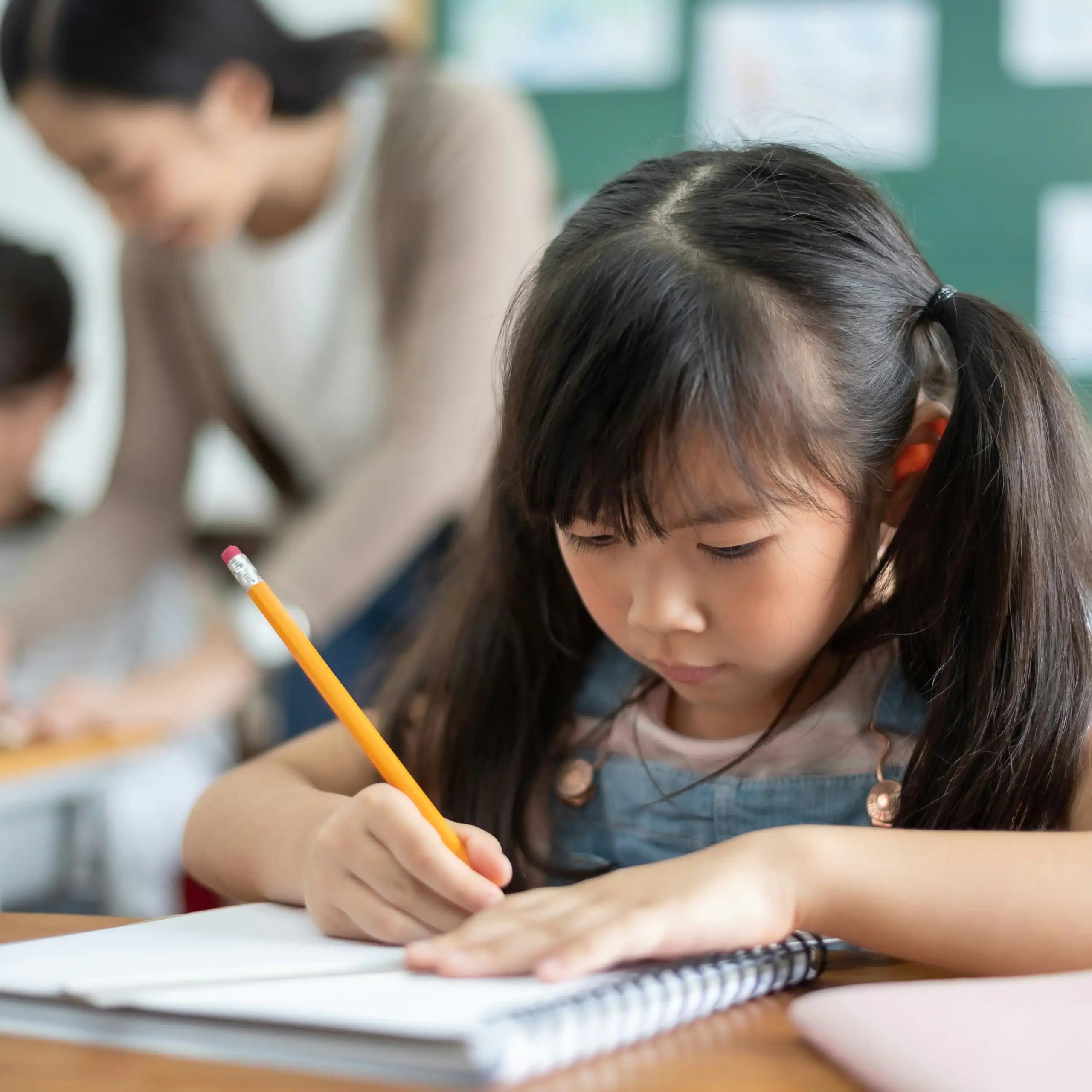 An Asian child in a classroom writing in a notebook with a pencil and behind her is a teacher in blurred focus with a student.