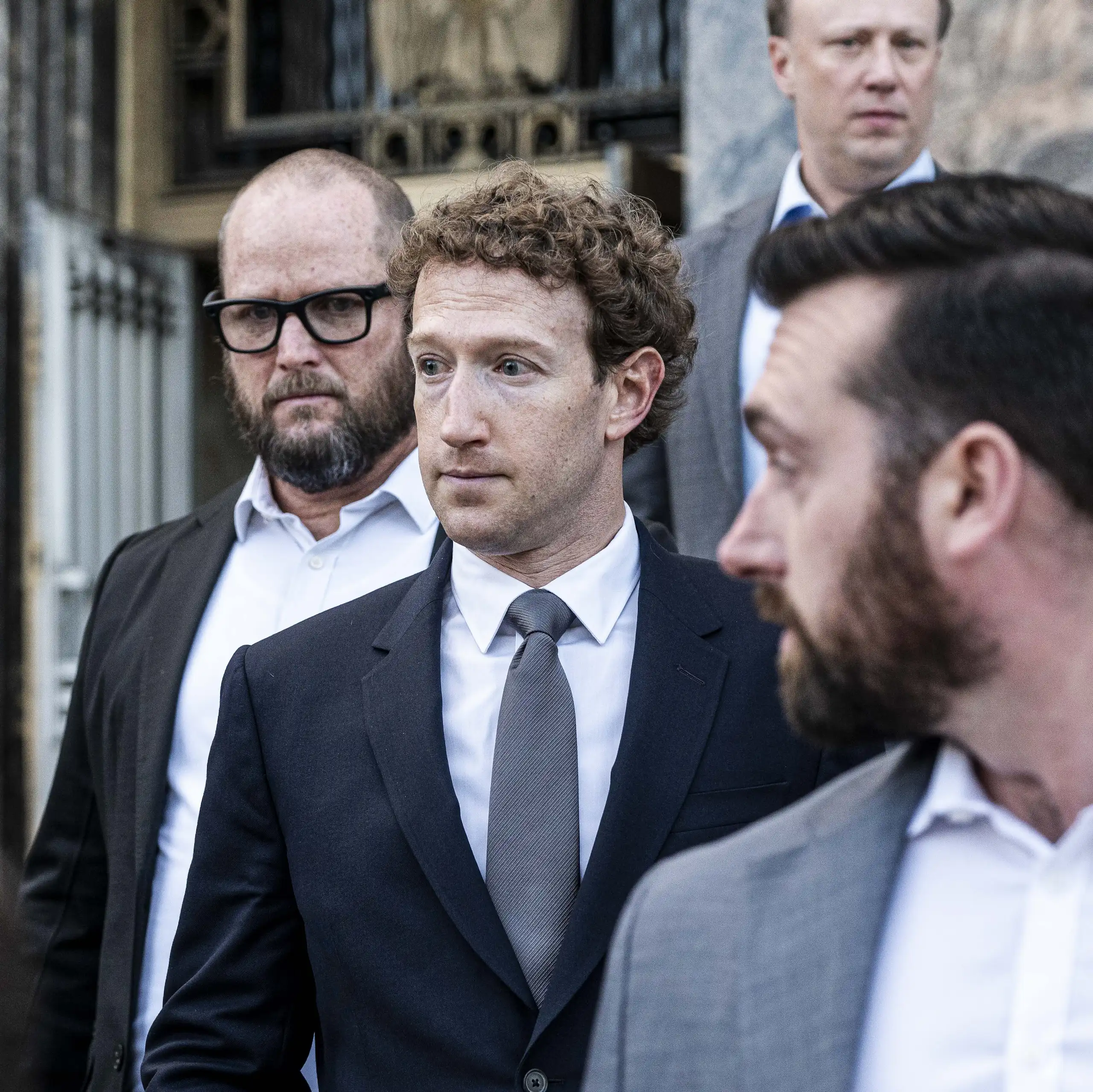 a man with curly hair wearing a business suit and tie surrounded by several other men outside a stone building