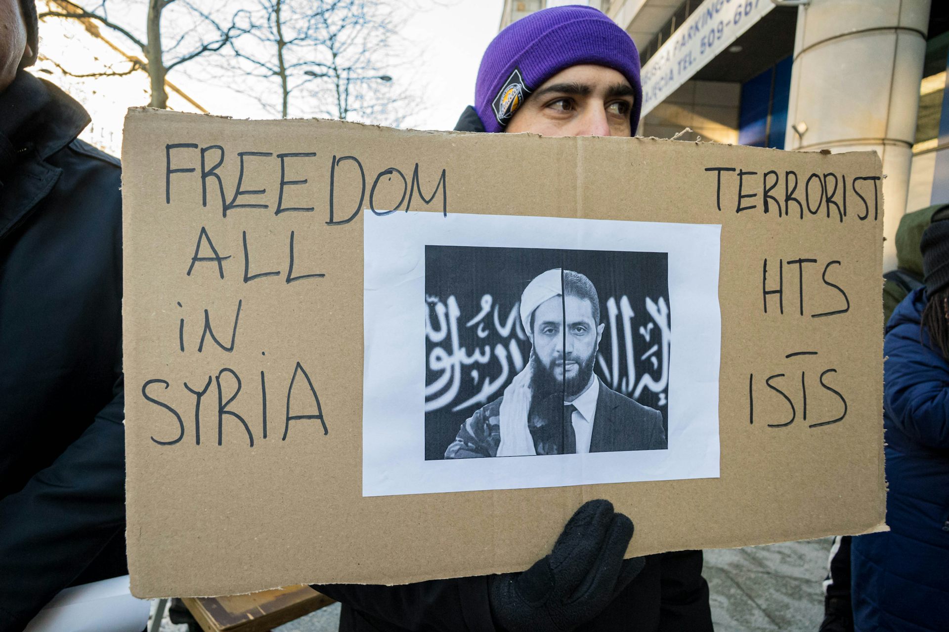 A man holds a cardboard banner with a man's face and the words 'Terrorist' on it.