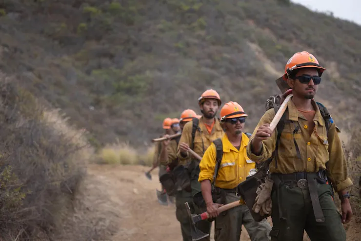 A line of firefighters in soot- and dirt-stained yellow jackets walk along a road carrying equipment.