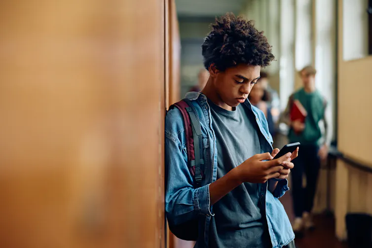 a teen boy leans against a wall while looking at a phone