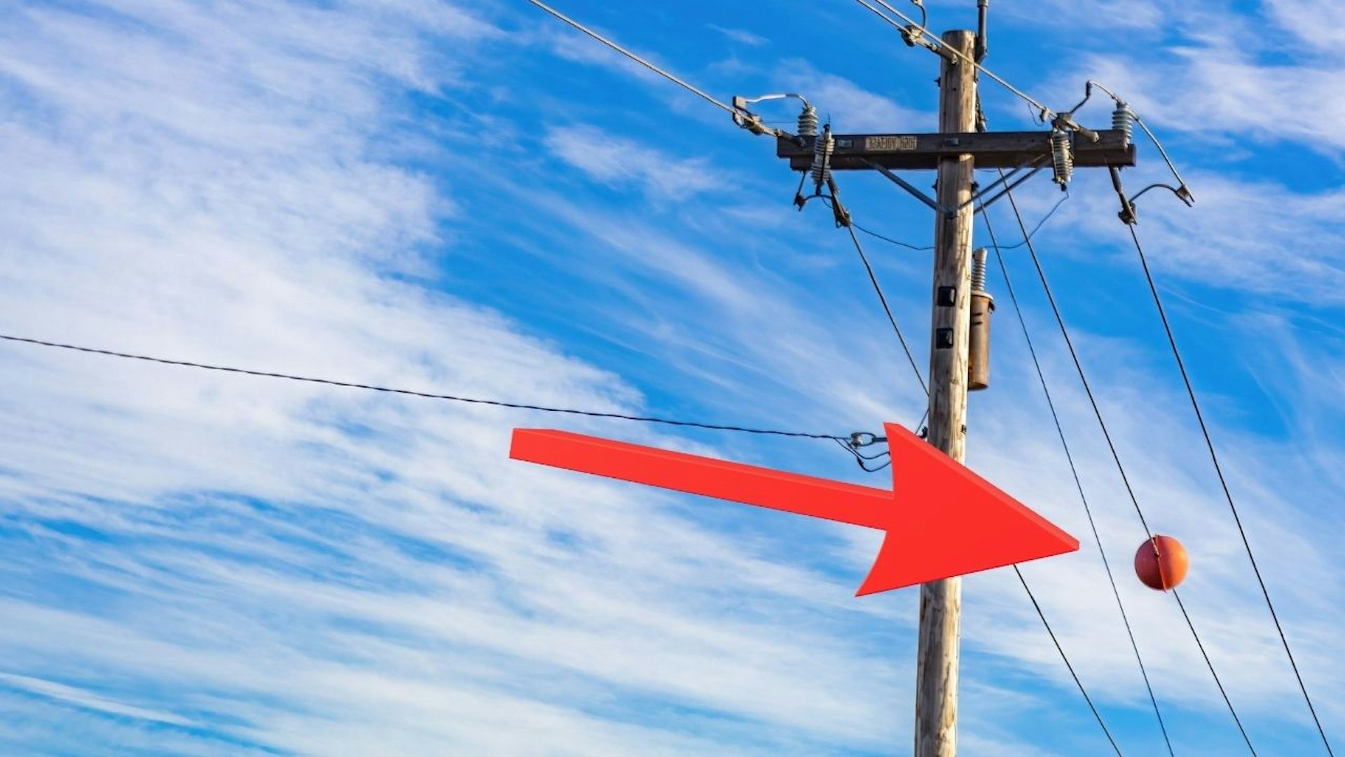 Power lines on tower. Orange ball hangs from one of the power lines There is an arrow pointing to the ball. The sky is bright blue with clouds.