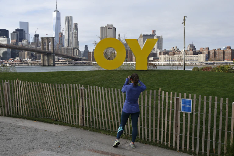 A woman in blue running clothes snaps a picture of a large yellow sculpture in a park by a river.