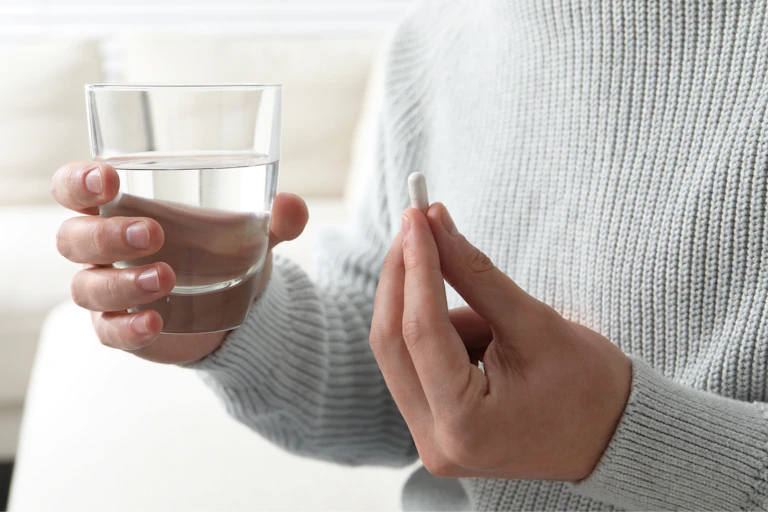 Close-up of a person's hands holding a glass of water and pill on blurred background.