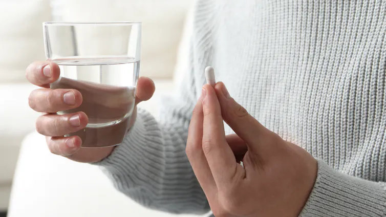 Close-up of a person's hands holding a glass of water and pill on blurred background.