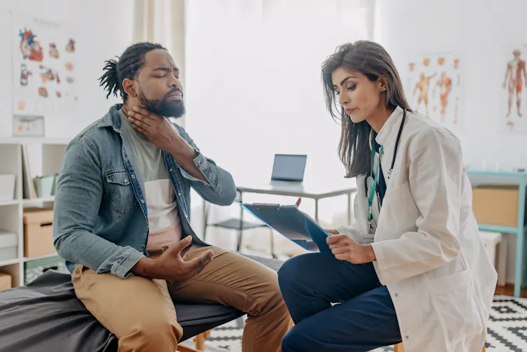 A patient sits on an exam table holding his throat while a doctor takes notes