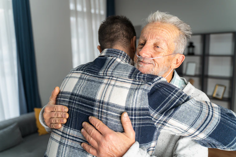 Older man with oxygen tubes in nose embraces son.