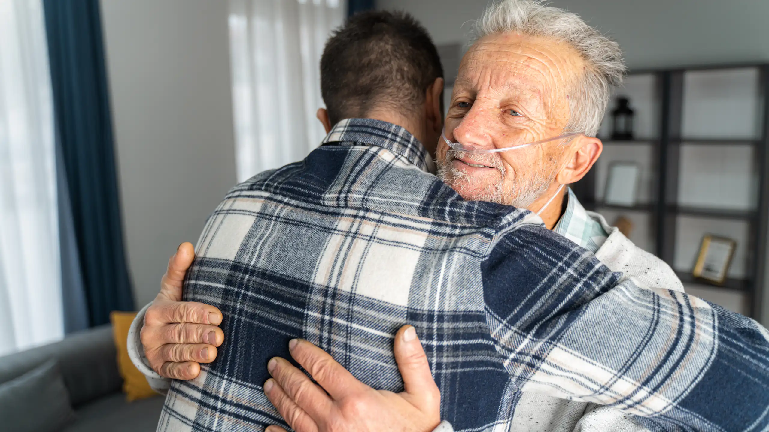 Older man with oxygen tubes in nose embraces son.