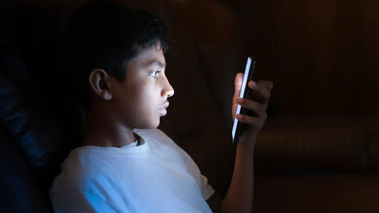 A young boy looks at a mobile phone in a dark room.