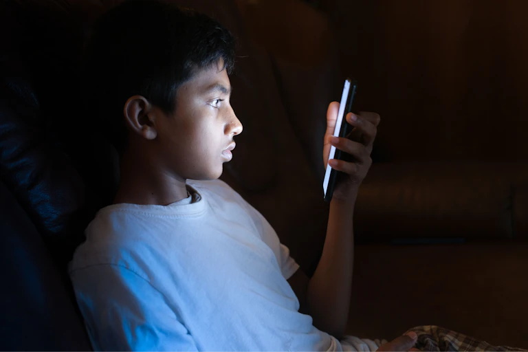A young boy looks at a mobile phone in a dark room.