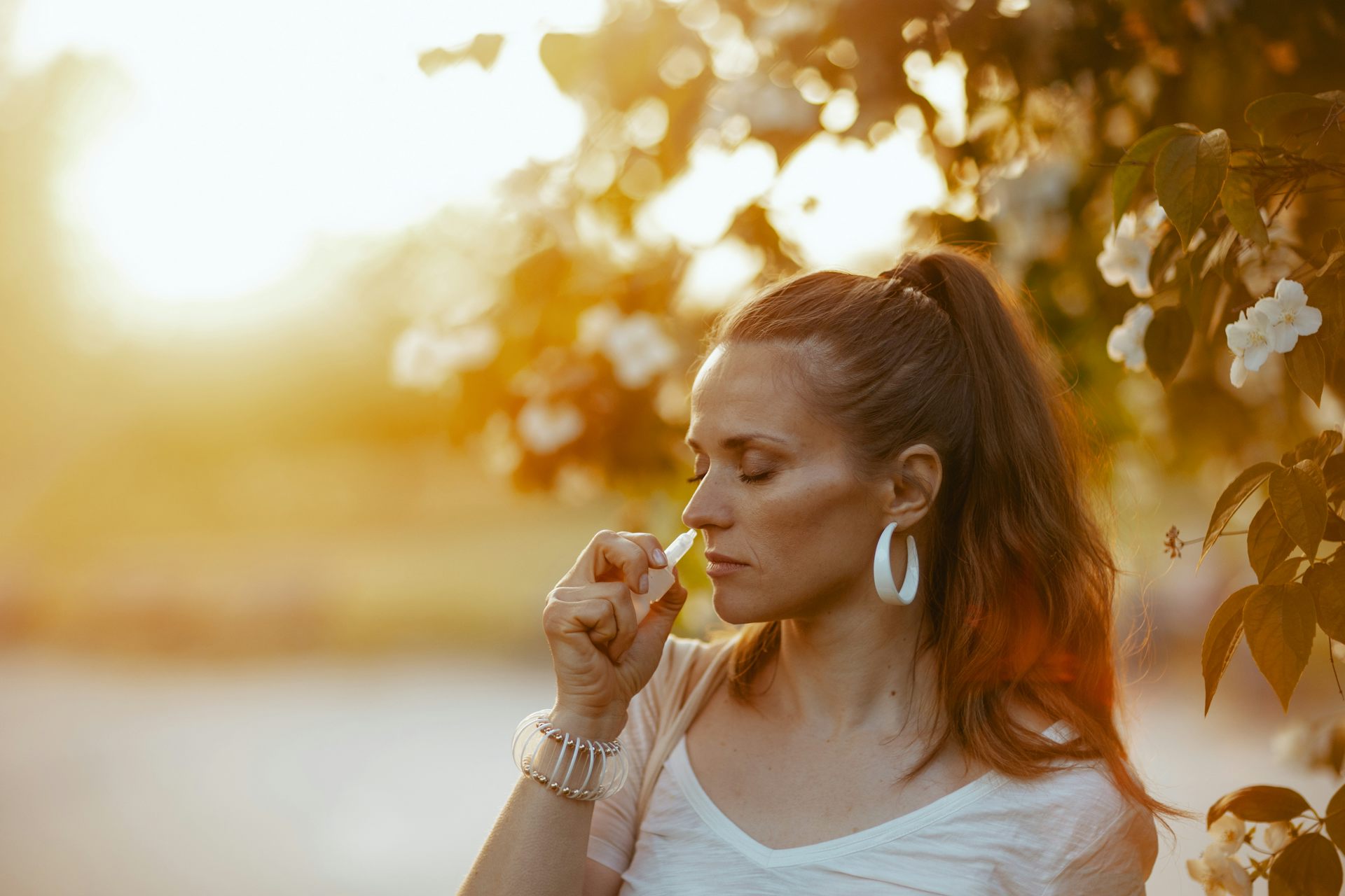Photo d’une jeune femme pulvérisant un produit dans son nez, sur fond ensoleillé.