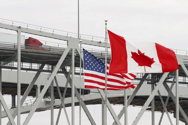 Two flags – one red and white with a red maple leaf, the other with white stars on a blue background and red and white stripes – wave next to a bridge with cars on it.