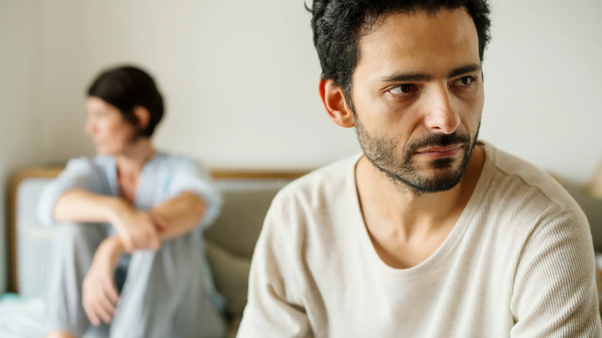 A distressed-looking man looks away while a woman in the background is sitting on a bed looking in the other direction.