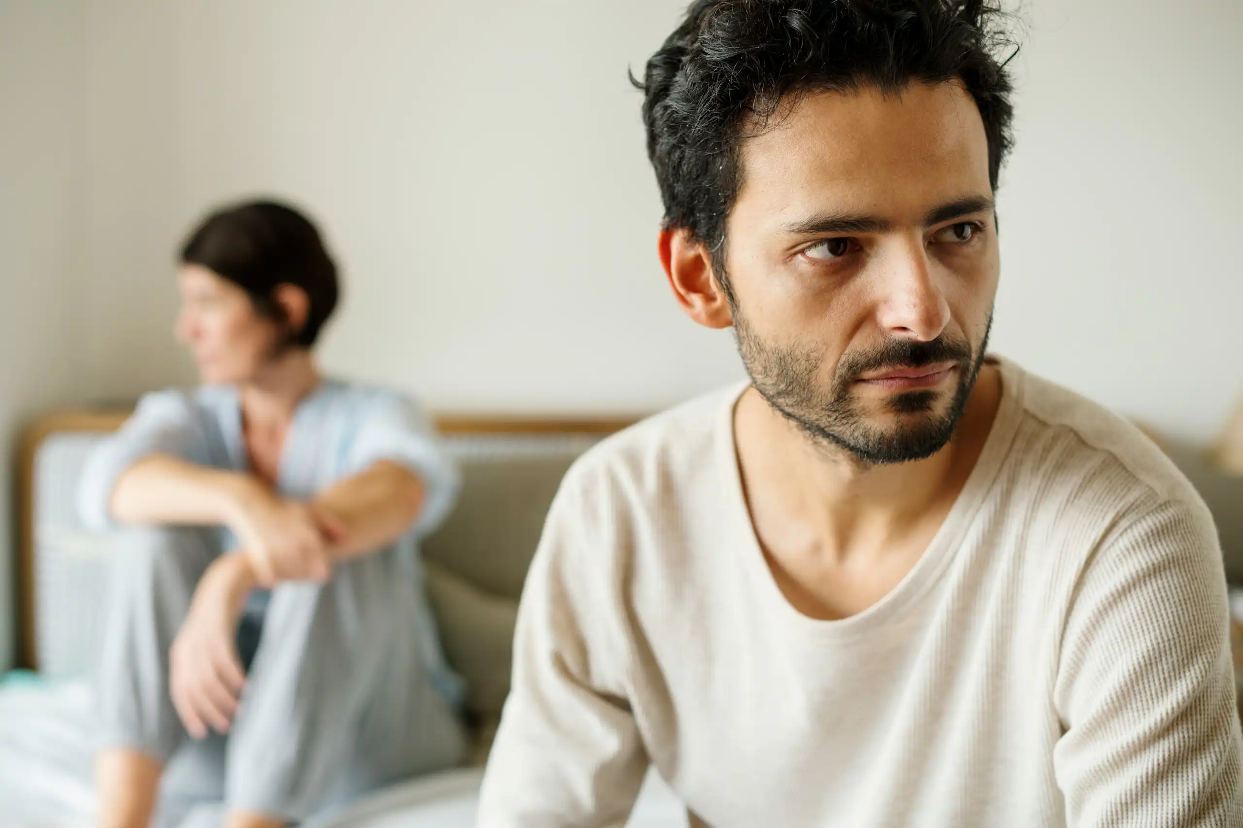 A distressed-looking man looks away while a woman in the background is sitting on a bed looking in the other direction.