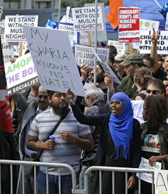 A woman holds a sign that reads, 'My Sharia Knows No Hate,' while standing behind a barricade with other protestors holding different signs.
