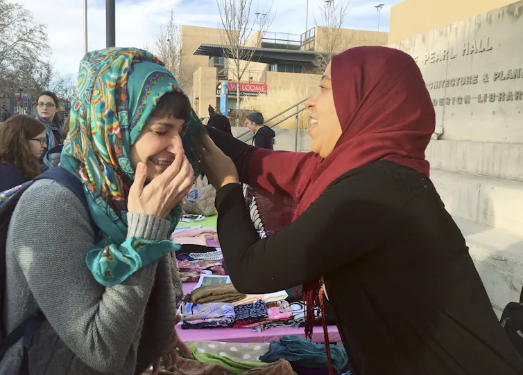 A woman in a red hijab helps another woman adjust a floral-print hijab.