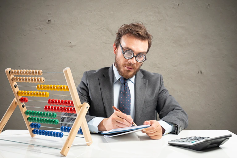 Man wearing glasses in suit sitting at desk holding pen and paper in front of abacus