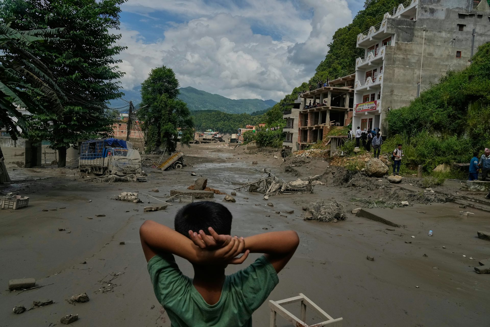 A boy photographed from behind with his hands behind his head looks at a vast expanse of mud and rubble.