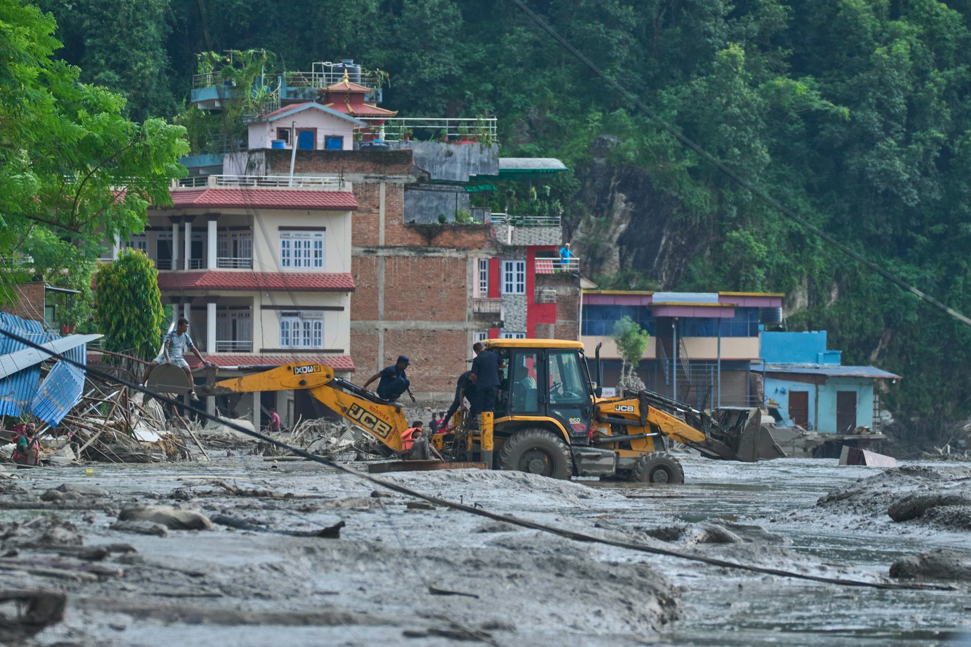 A large piece of equipment like a bulldozer digs in a large expanse of mud with houses in the background.