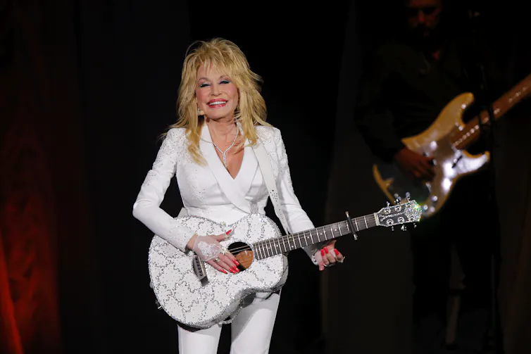 A blonde woman in a white suit smiles as she stands on stage, holding a white guitar.