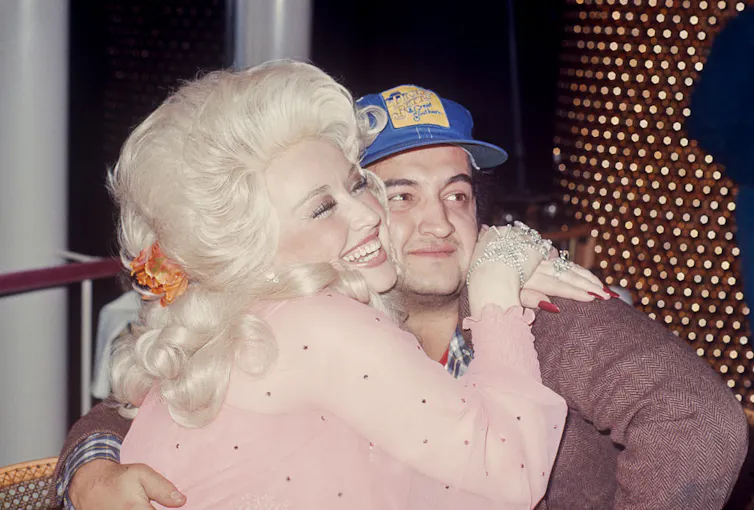 Young woman with blonde hair smiles while hugging a young man wearing a blue baseball cap.