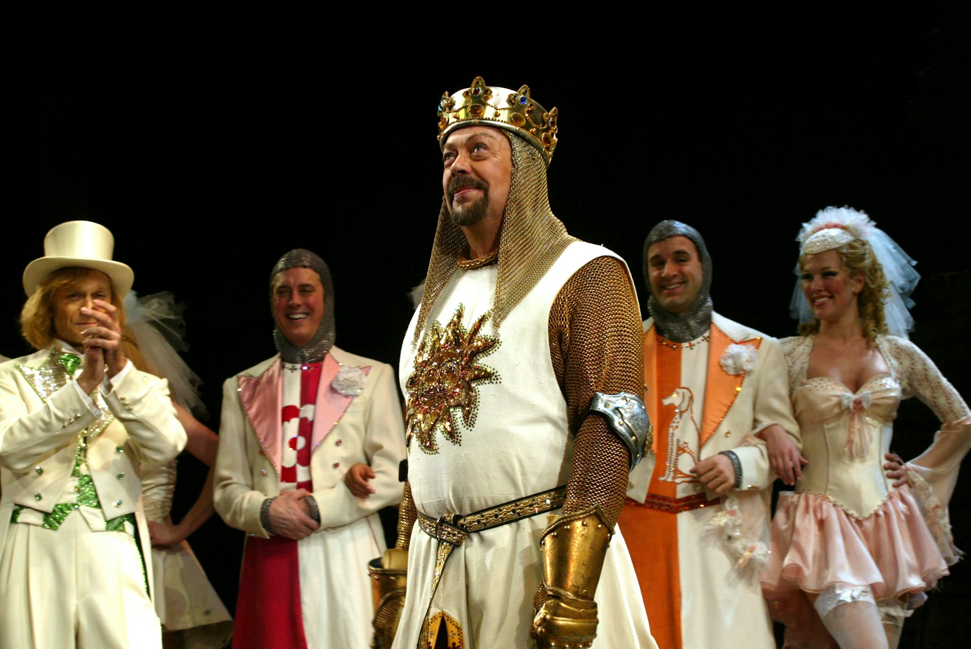 A middle-aged white man in a beard, dressed up like a medieval king, stands on a stage with other actors