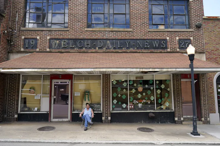 A woman sits outside a brick building with a sign reading 'Welch Daily News' across the top.