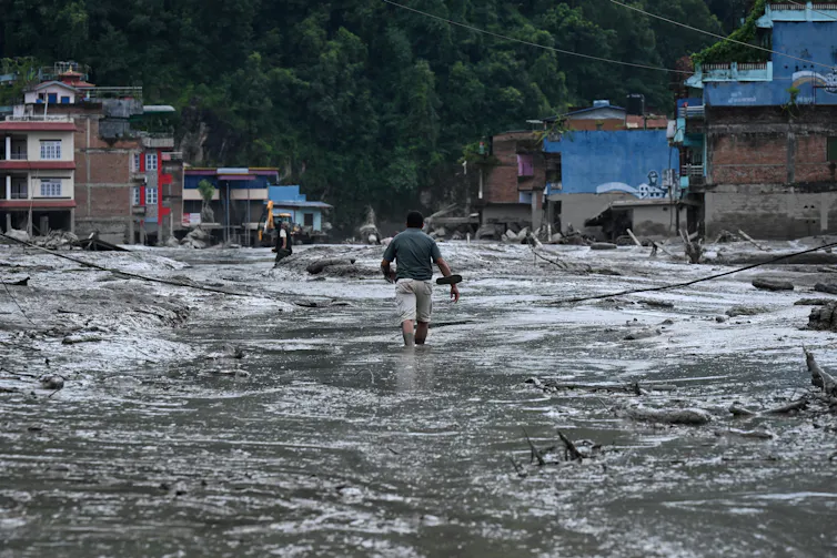 A man walking through thick mud, passed ruined houses.