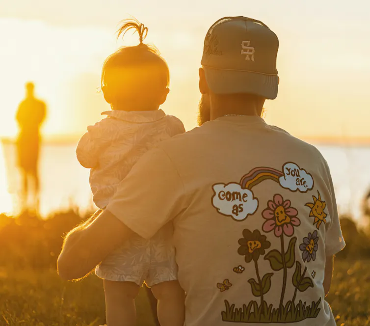 A man in a backwards cap and t-shirt with flowers and 'Come as you are' on it holds a small child as they watch the sea and sun setting.
