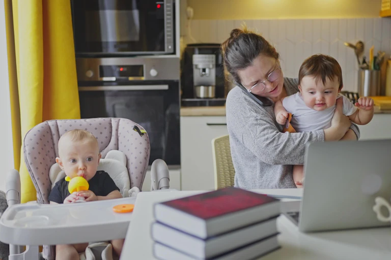 woman sitting at a table speaking on the phone and struggling with a baby and a young child in a high chair