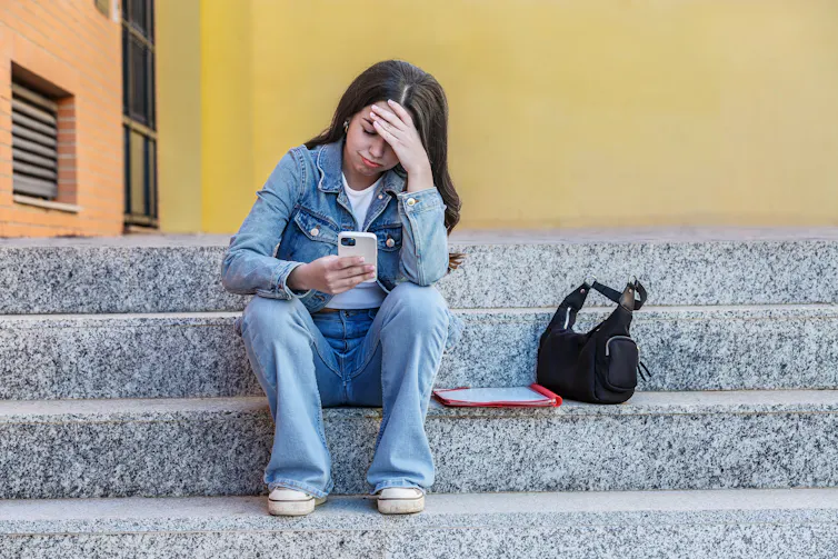 A young woman sits on a concrete stairway looking sad while gazing at her cellphone.