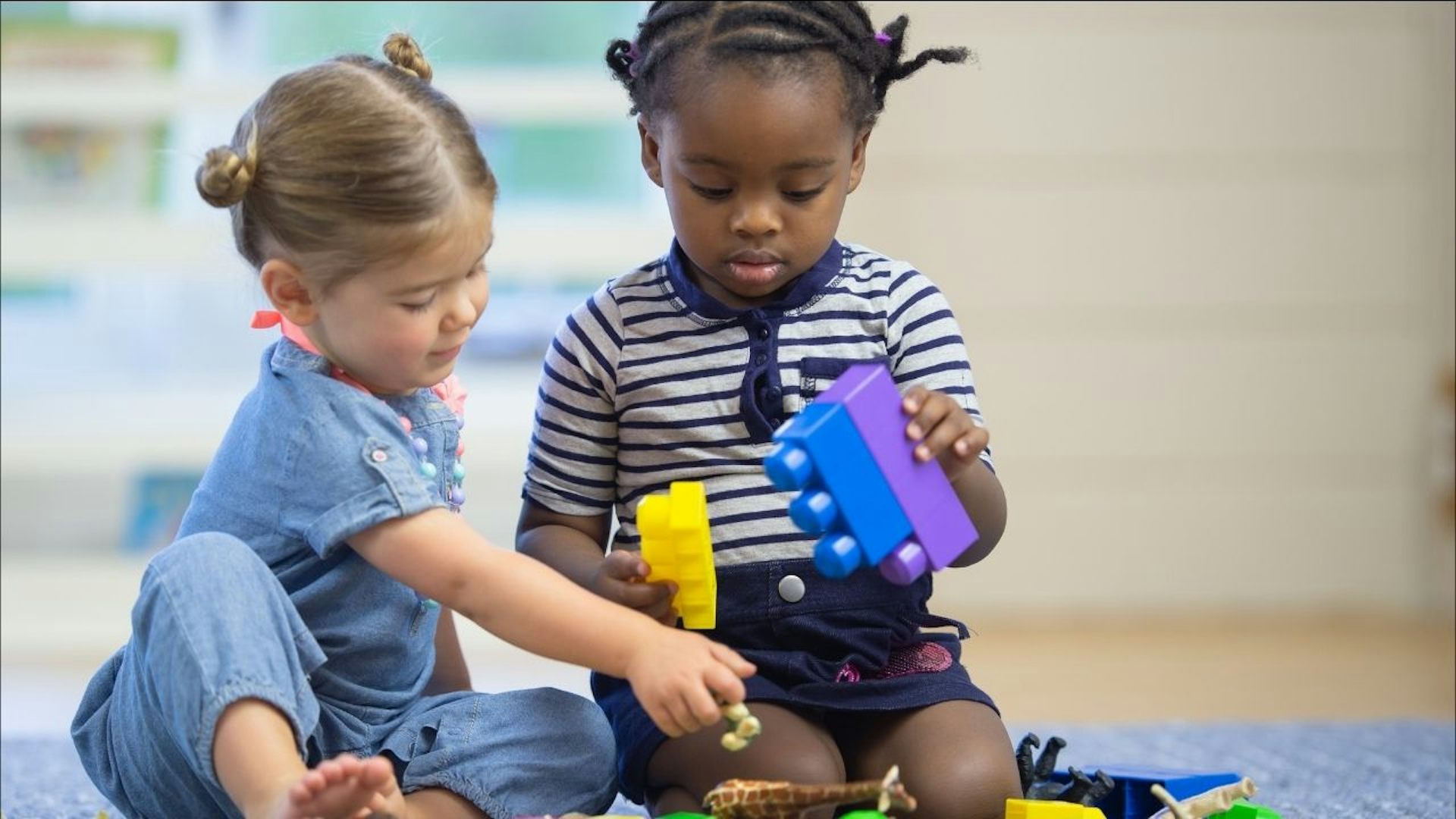A white girl and a Black girl play with colorful toys while sitting on the floor
