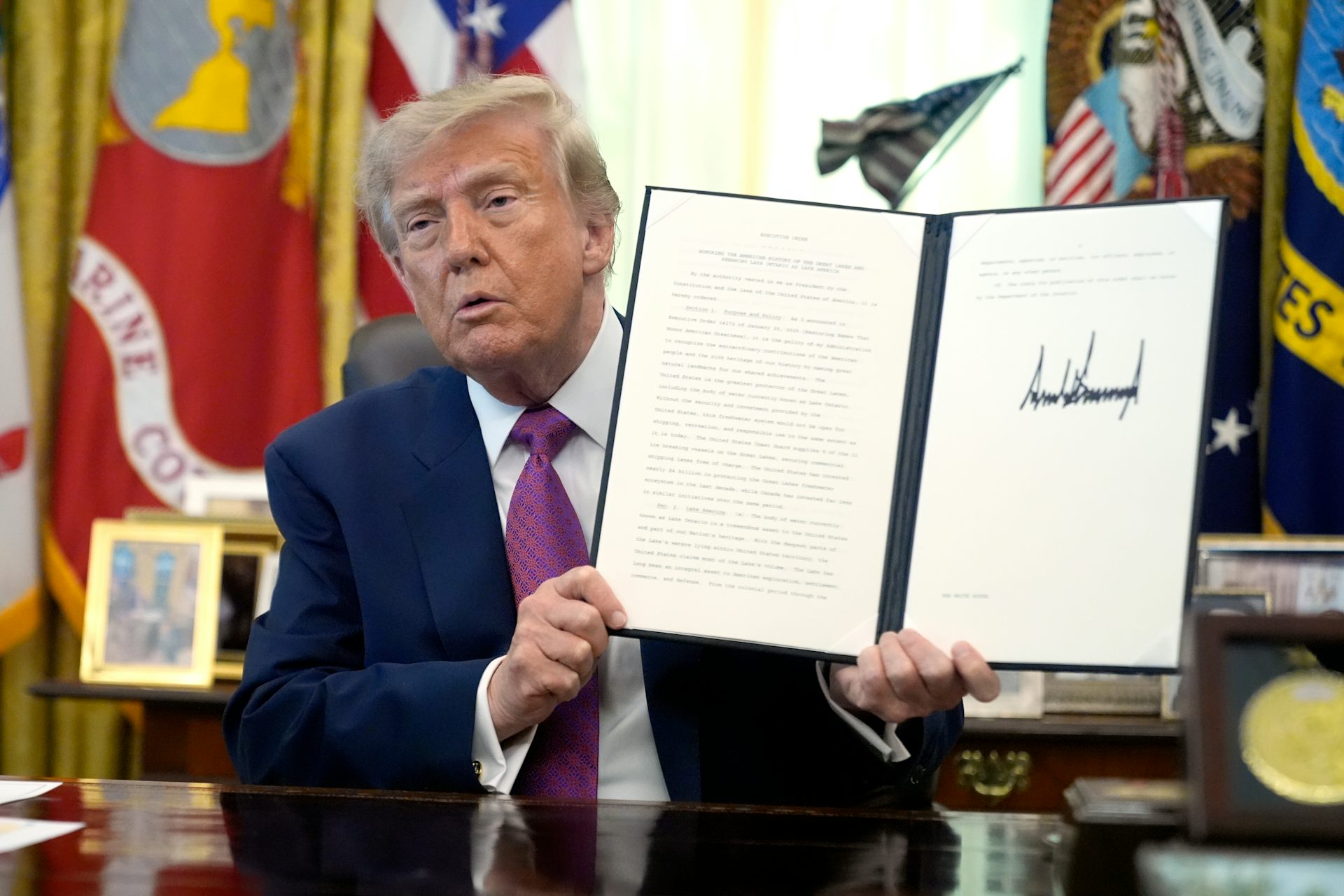 an old orange-hued man holds up a document with an unnecessarily large signature on it