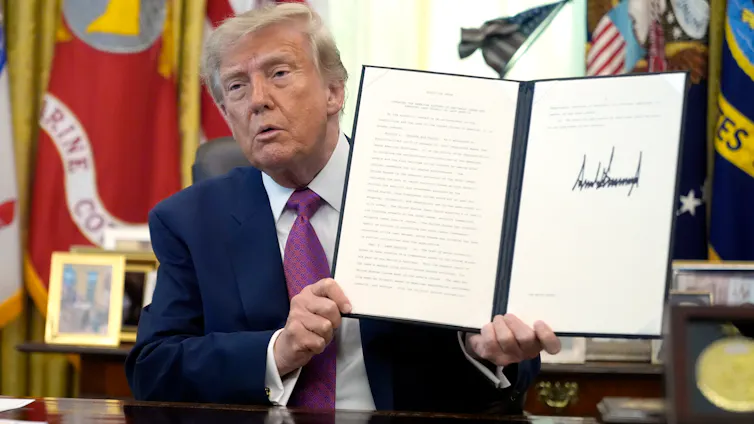 an older orange-hued man holds up a document with a large signature on it