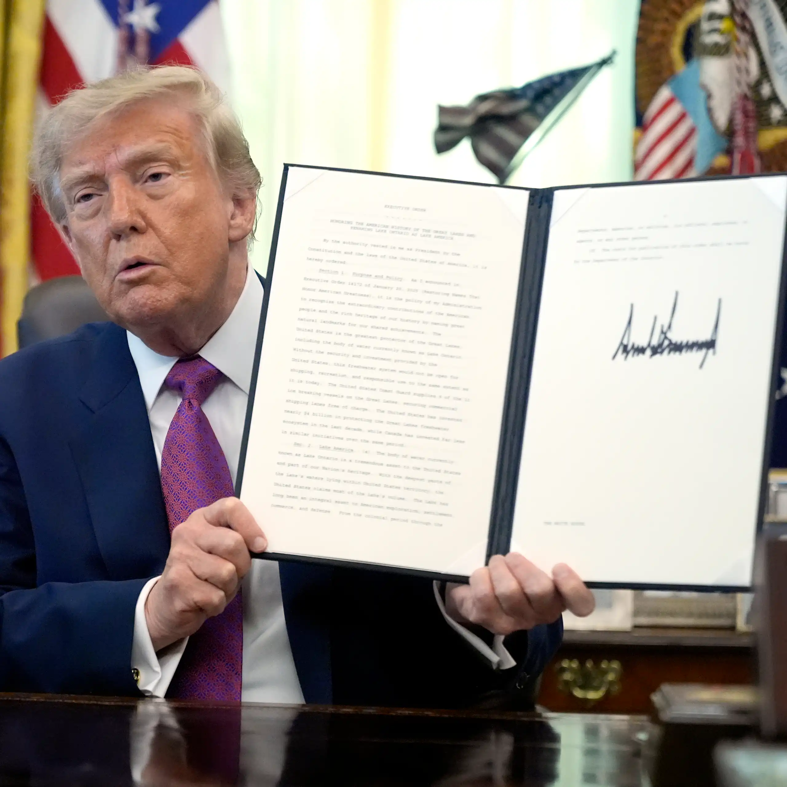 an old orange-hued man holds up a document with an unnecessarily large signature on it