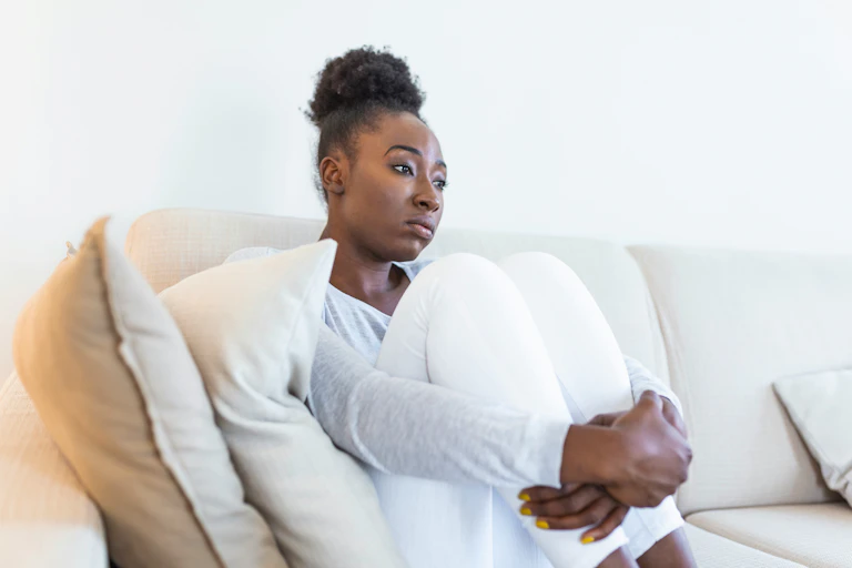 A woman looking pensive, sitting on a white sofa hugging her knees to her chest.