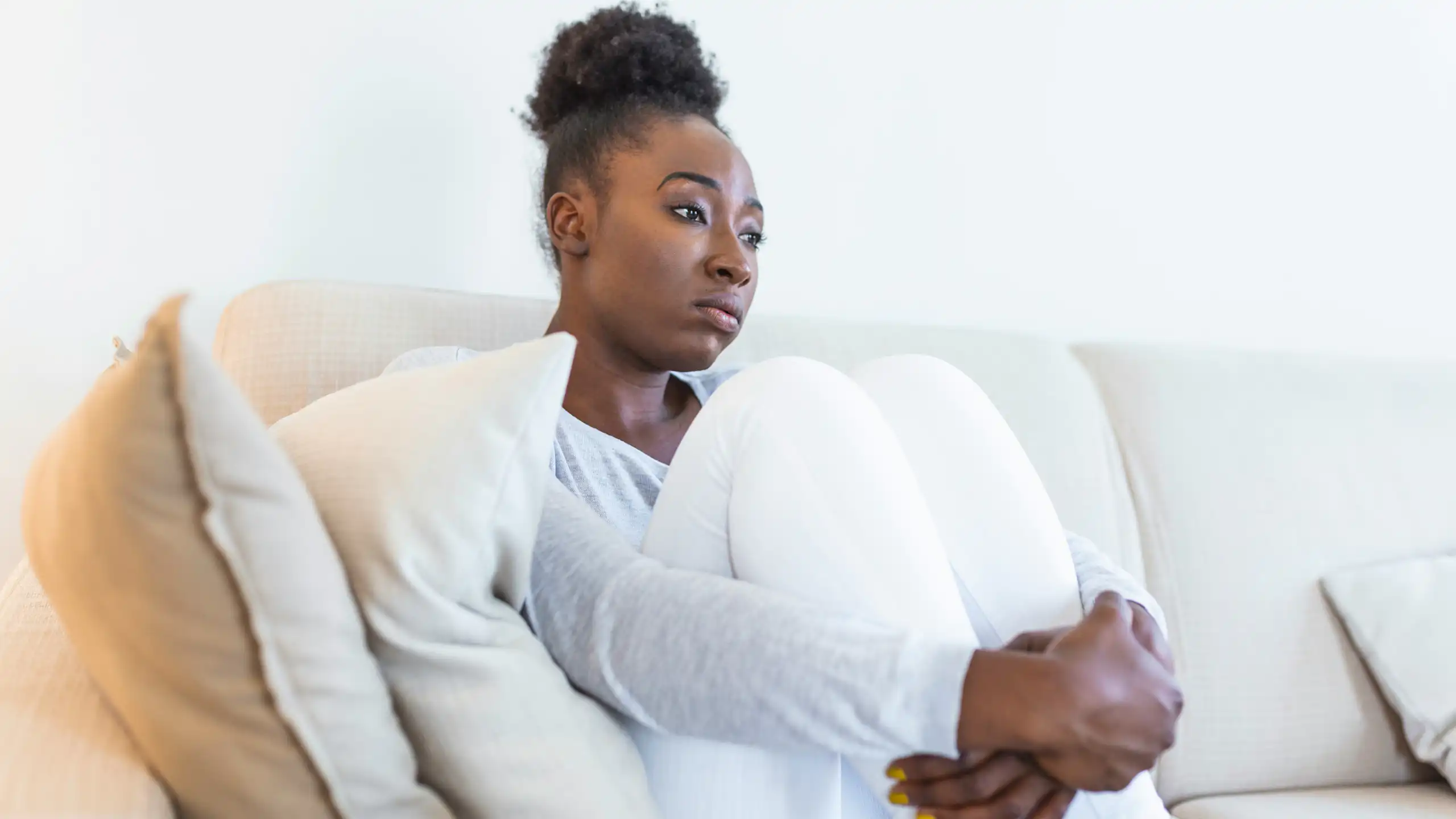 A woman looking pensive, sitting on a white sofa hugging her knees to her chest.