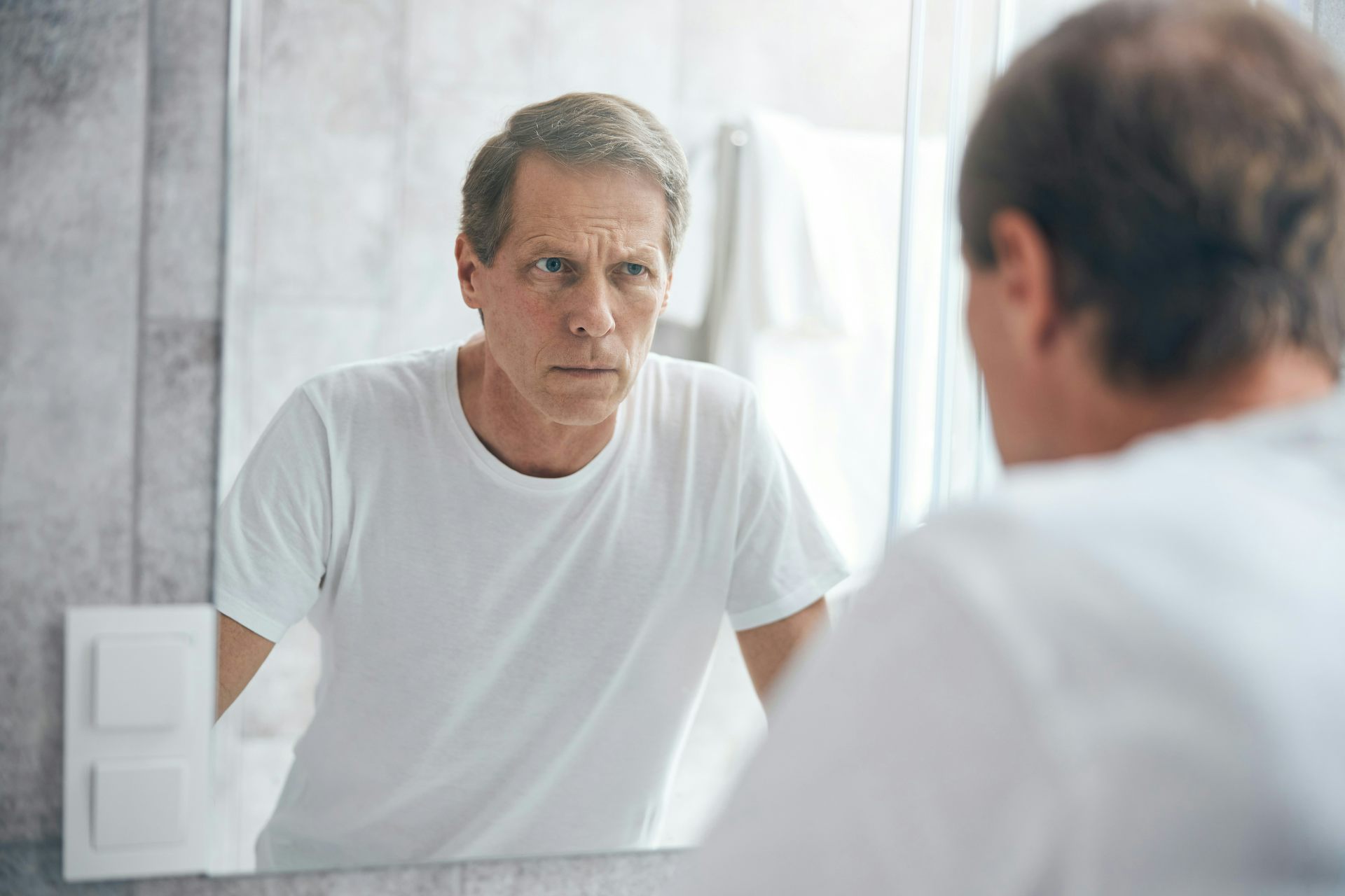 A man in a white T-shirt with grey hair looking in a mirror