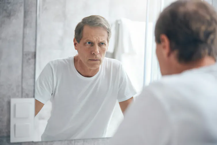 A man in a white T-shirt with grey hair looking in a mirror