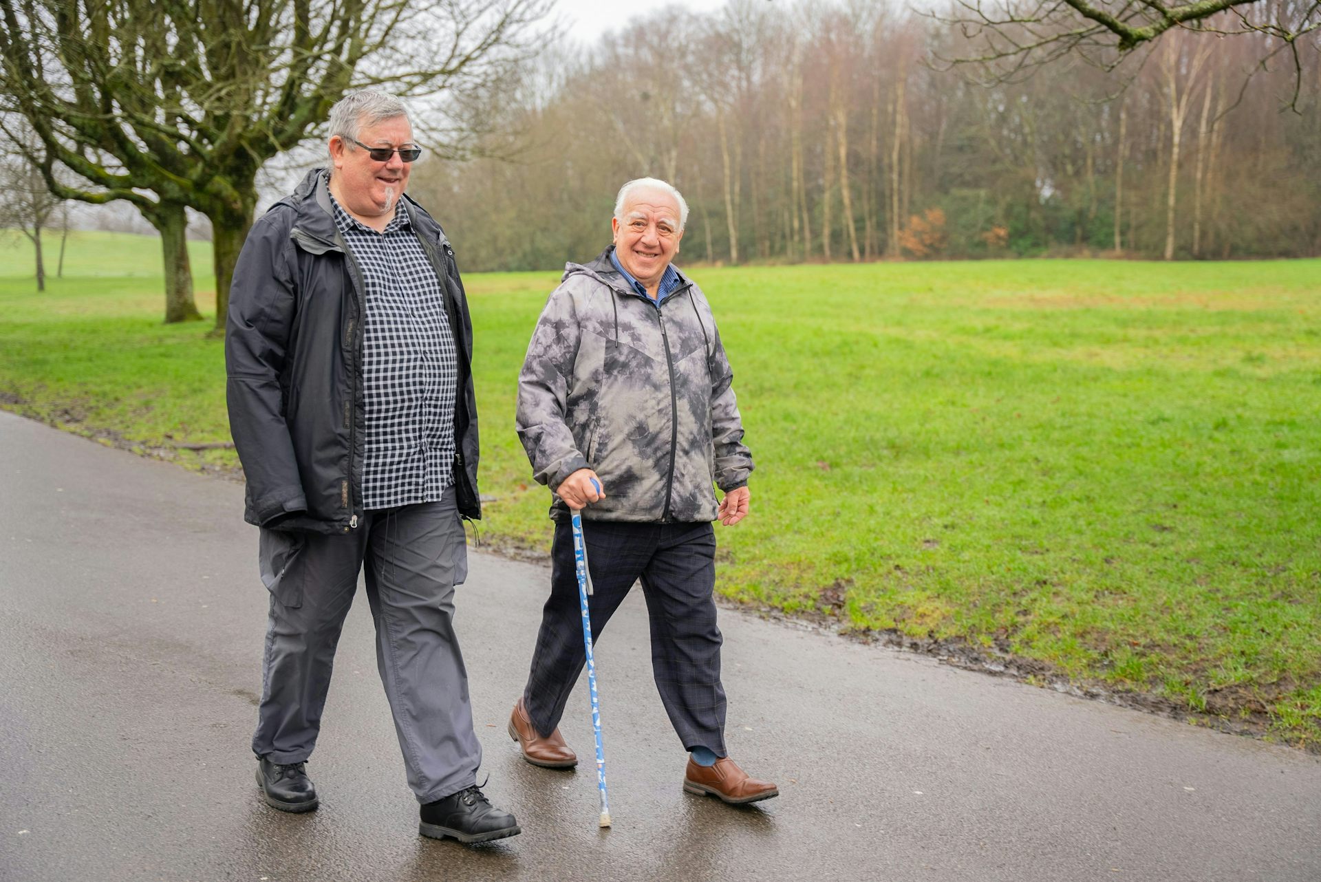Two men walking outdoors on a path; one of them uses a cane
